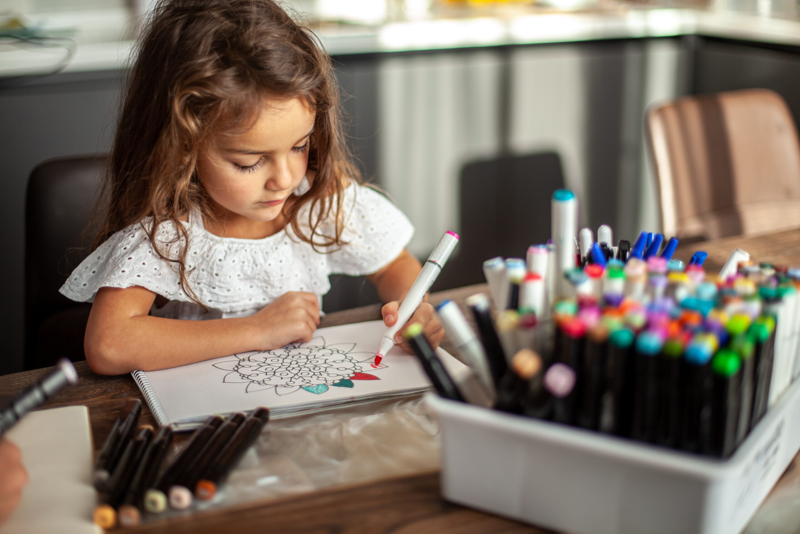 A little cute girl draws a circular mandala pattern in the album with art markers.